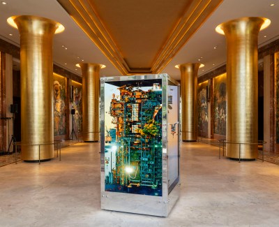 A six foot silver rectangular box with multicolored wires inside sits in a atrium of gold columns.