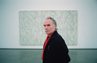 A white man in a black jacket and red scarf in front of an abstract painting with squiggles and lines.