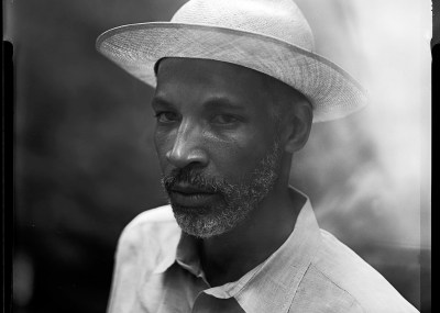 A Black man in a straw hat and a collared shirt looking at the camera.