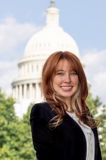 Kadison Willis, a woman with long hair wearing a black blazer and white blouse, smiles while looking directly into the camera. Behind her is the white dome of the Capitol building.