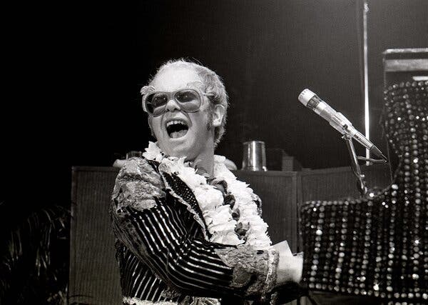 A black and white photograph from 1974 of Elton John, at a piano with a microphone, in a shiny stripe and ruffled top.