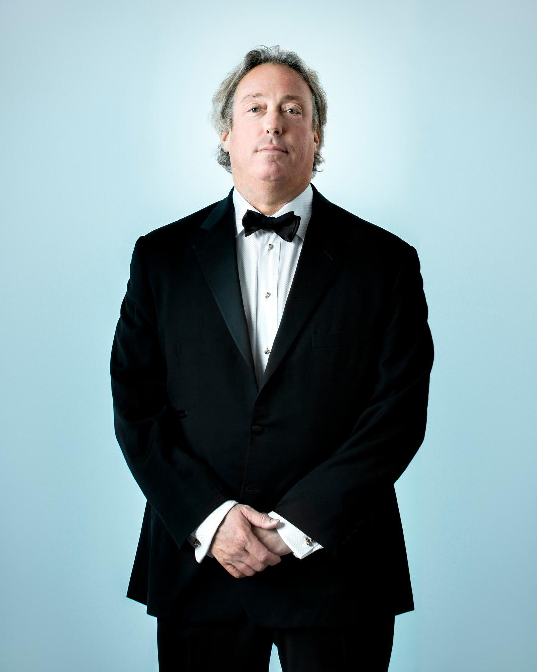 A man in a tuxedo poses for a photo in front of a light blue background