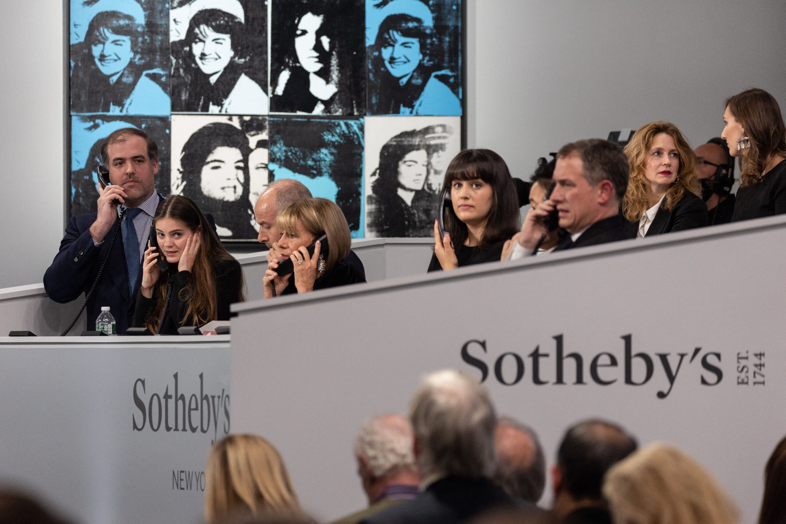 People speak to bidders on the phone, standing in front of an Andy Warhol print