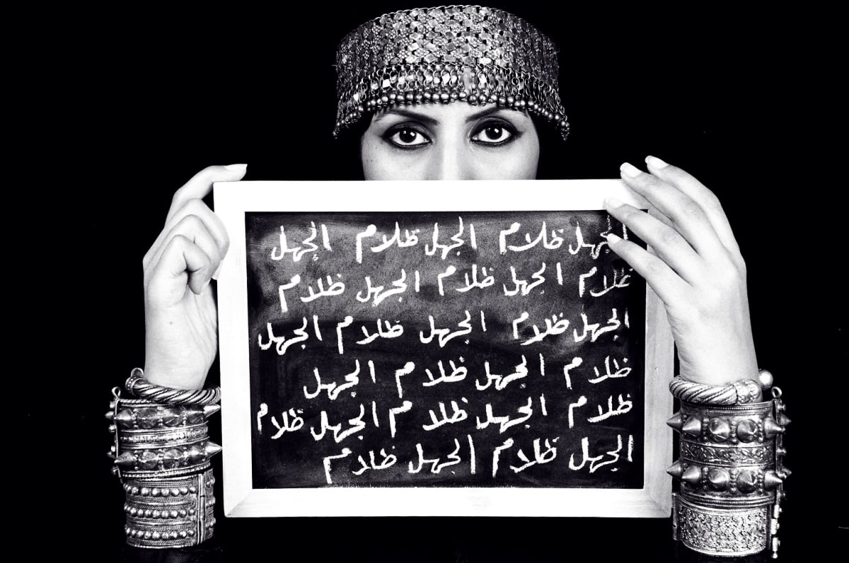 A black and white photo of a woman holding a slate on which there is Arabic writing