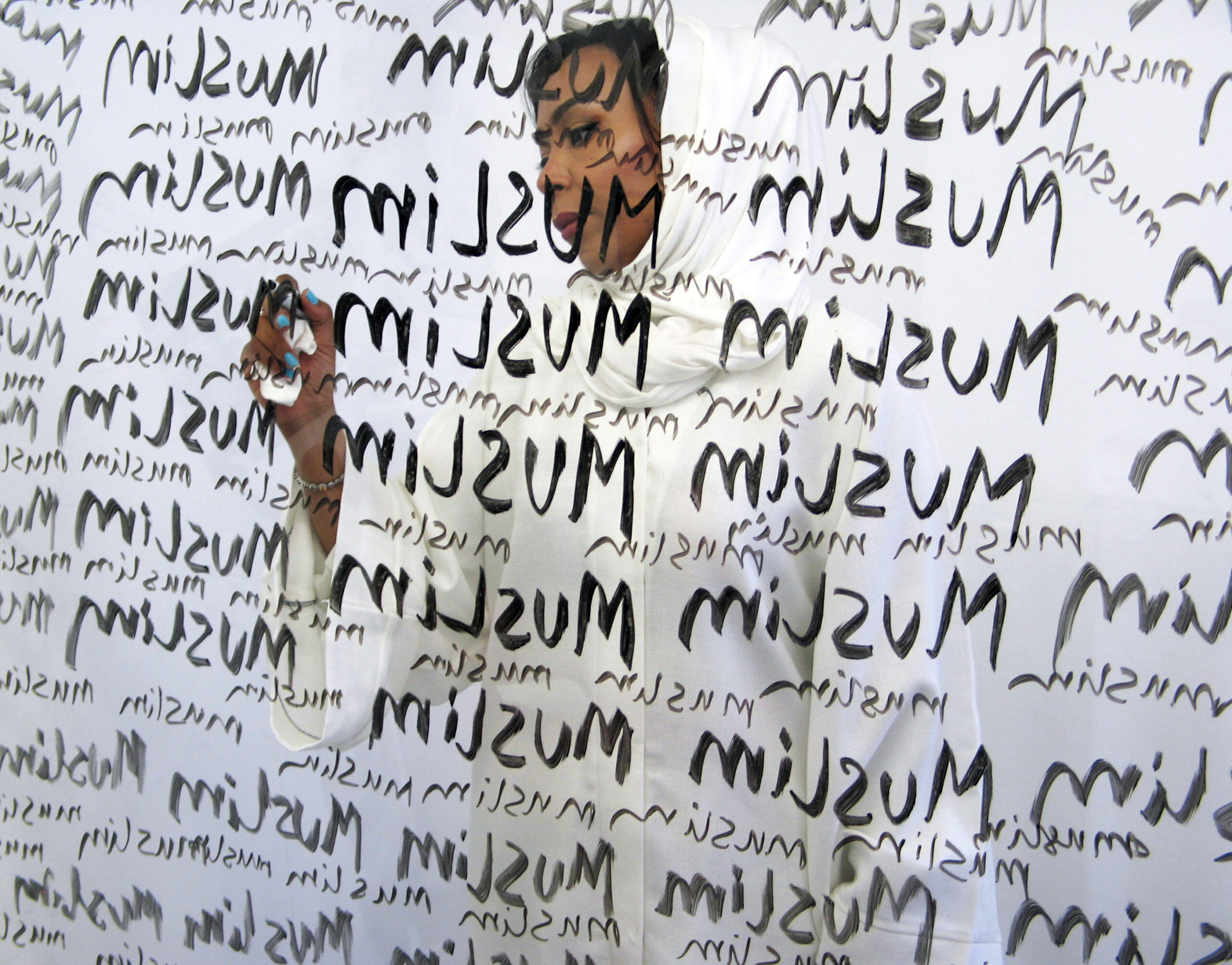 A man writes the word 'Muslim' repeatedly on a piece of glass using black marker