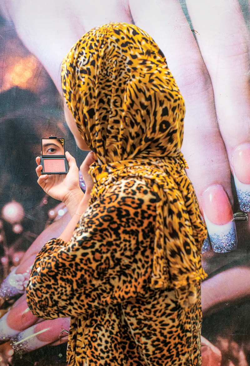 A woman in a leopard print headscarf stands turned away from the camera holding a compact