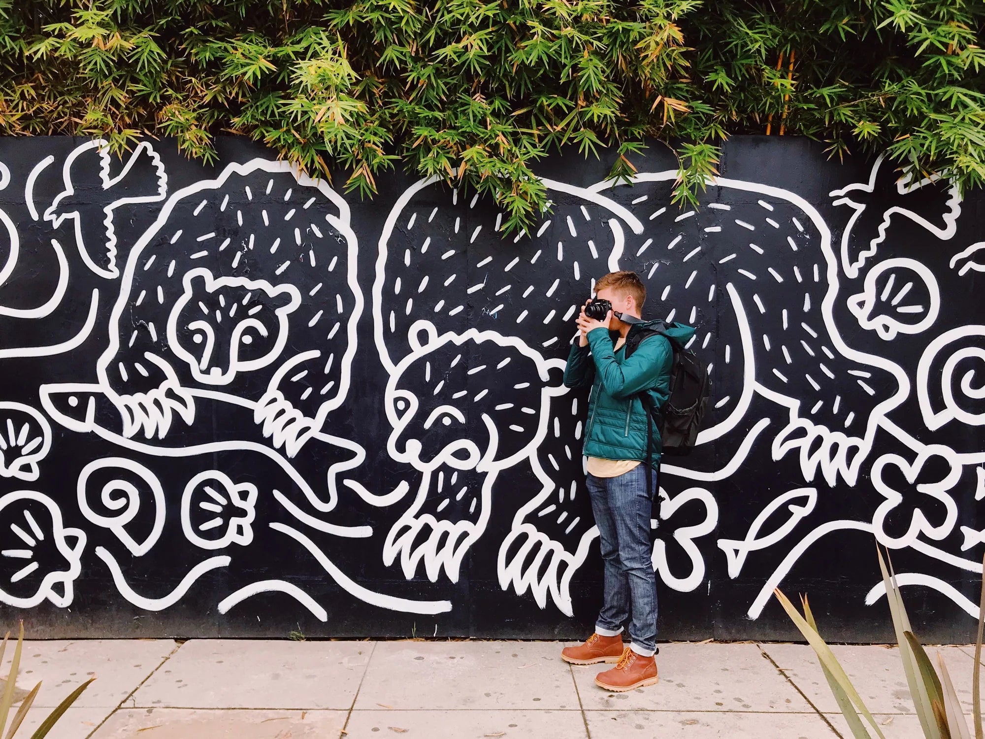 A man stands in front of a large street art mural taking a photo