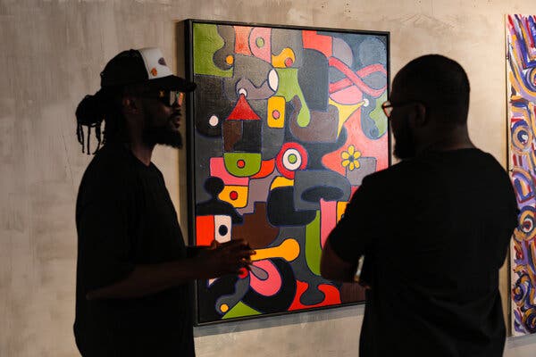 Two men, one wearing a baseball cap and sunglasses, seen from behind, stand in front of a brightly colored abstract painting.