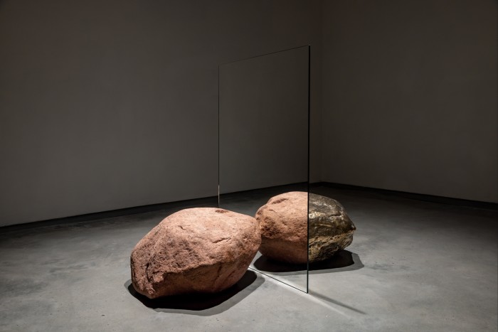 A large granite rock sits in front of a mirror at an angle; behind the mirror, part of a golden rock completing the mirror image is visible