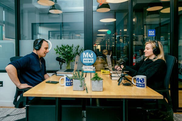 Mosheh Oinounou, left, wearing a dark shirt and headphones, sits across a table from Jill Wagner, who is wearing a dark sweater and headphones and typing on a laptop. On the table are two mugs that say “Mo News” along with a round Mo News sign at the far end of the table.