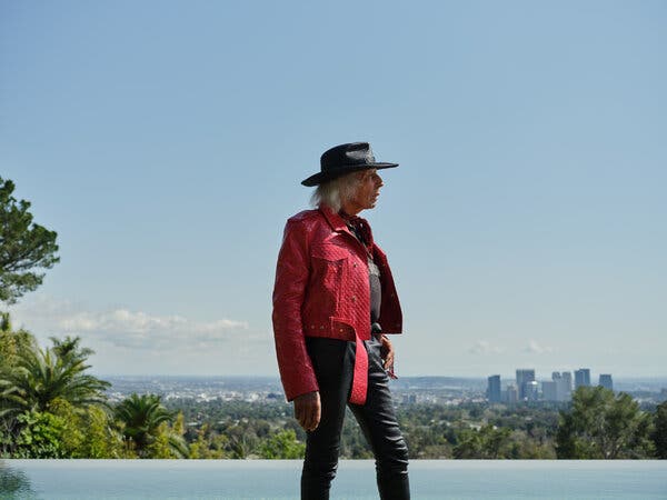 A frizzy-haired man in a western hat and a red jacket and black pants walks by an infinity pool.
