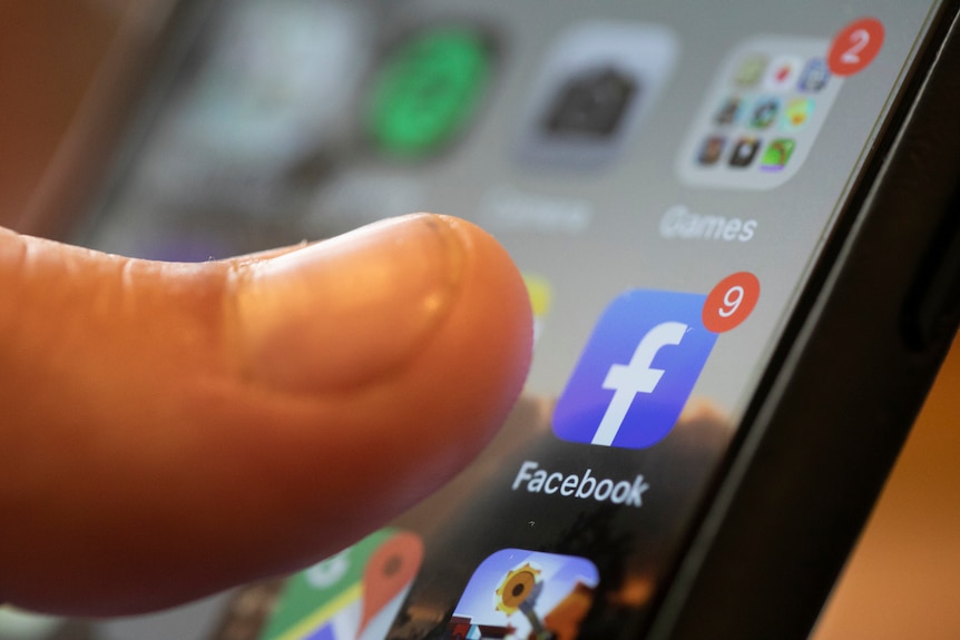 A person's thumb edges towards a Facebook app icon on a smartphone. 