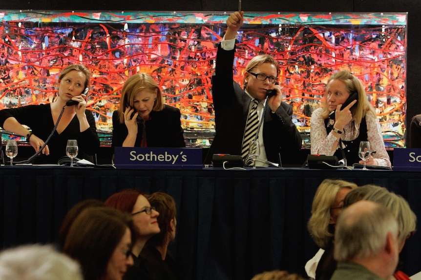 Four women and one man sit at a long table talking on the phone. The man, in the centre, has his right hand raised to make a bid