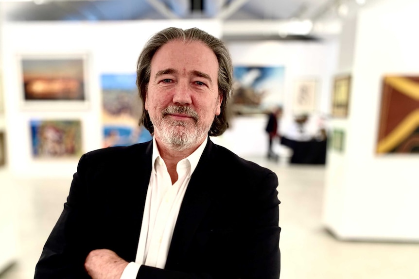A man wearing a white shirt and black jacket stands in front of artworks