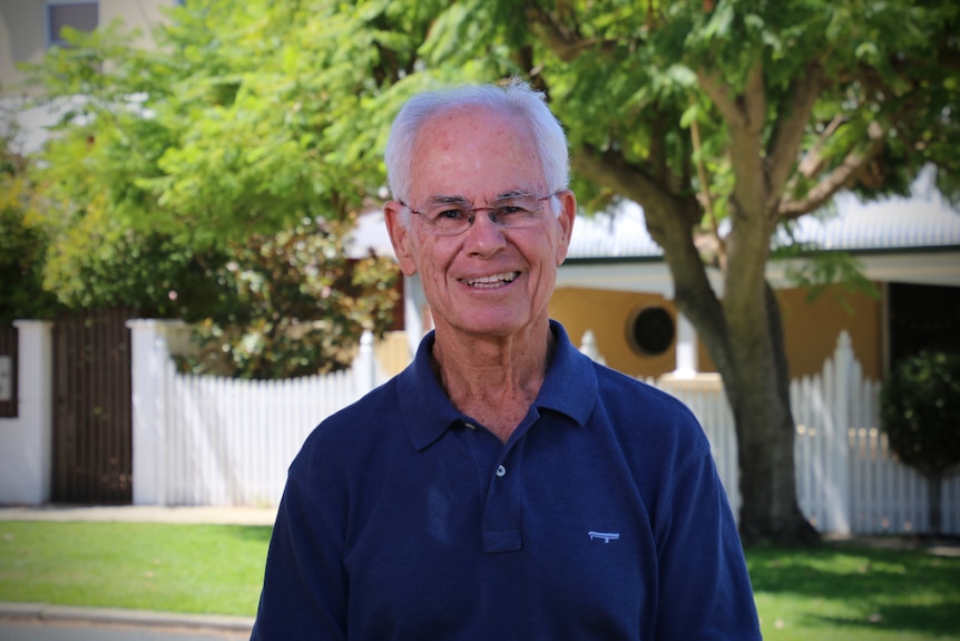 A man in a blue polo shirt smiling in front of a house with a white picket fence