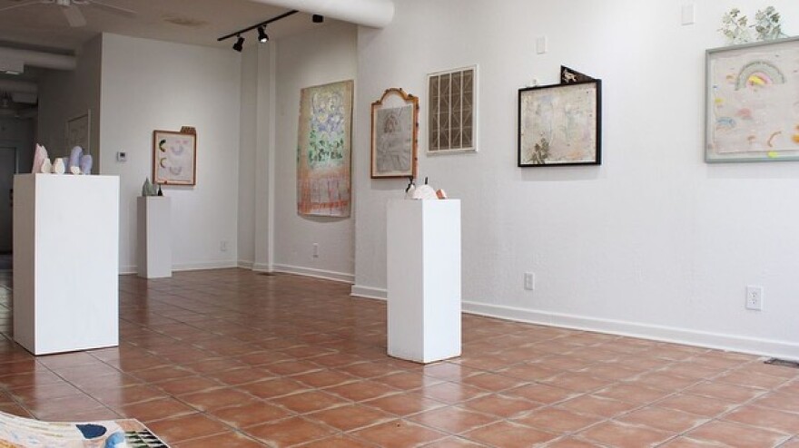 A white walled gallery space with brown tiled floors. Paintings are on the walls and there are three pedestals  displaying small sculptures. 