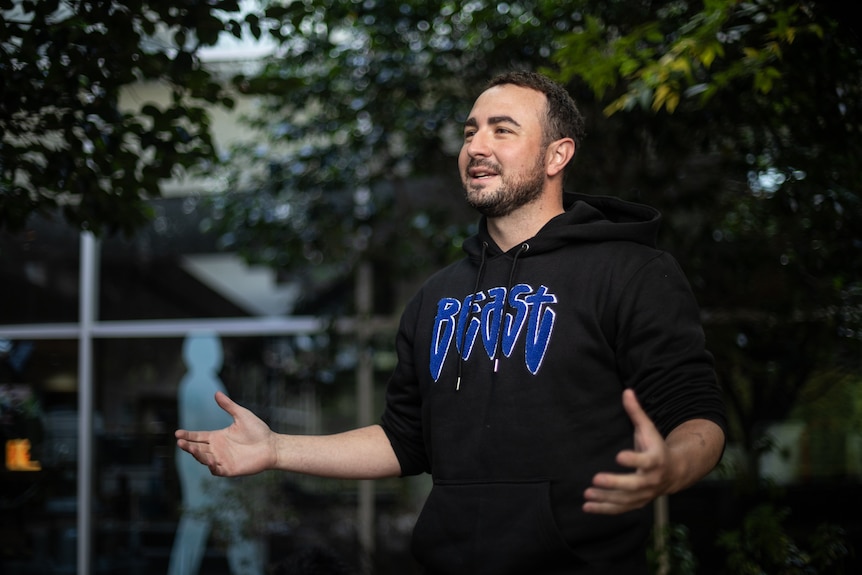 Man in black hoodie speaks passionately, his hands oustretched