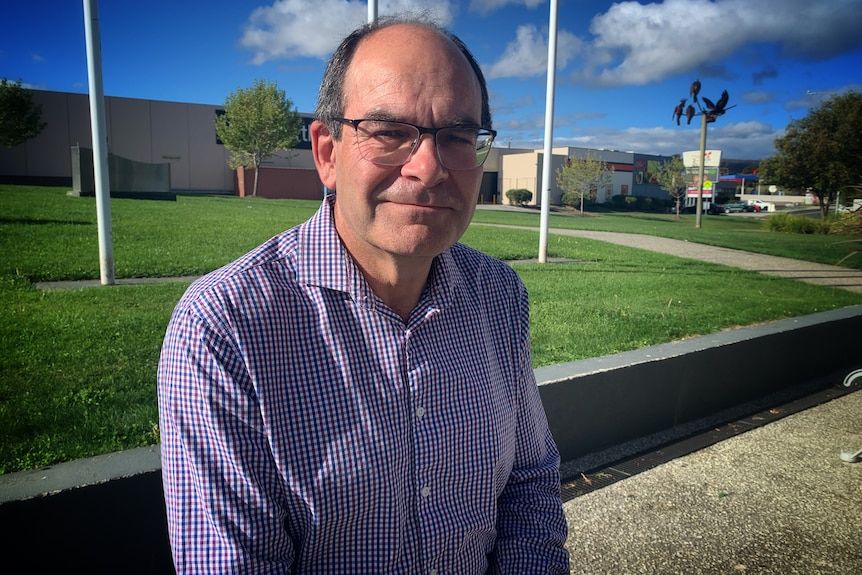 A seated man looks at the camera, with a lawn and shopping centre in the background.