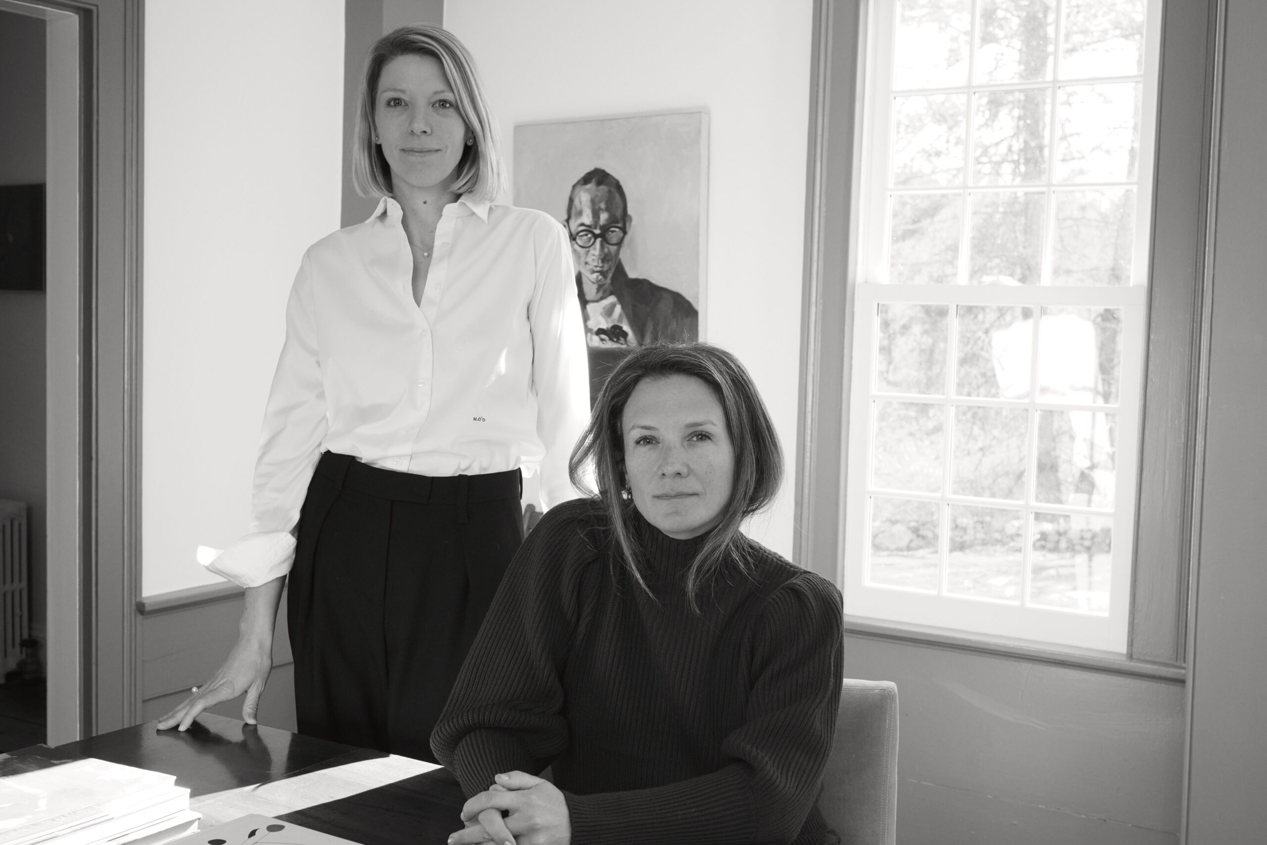 A black and white photo of two women in an office: one in a white shirt and black skirt standing and one in a dark sweater seated with her hands clasped