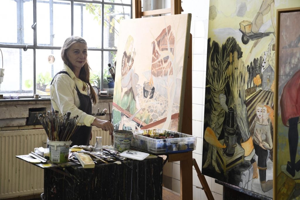 Rosa Loy, a middle aged white woman with long hair stands at an easel in an art studio.