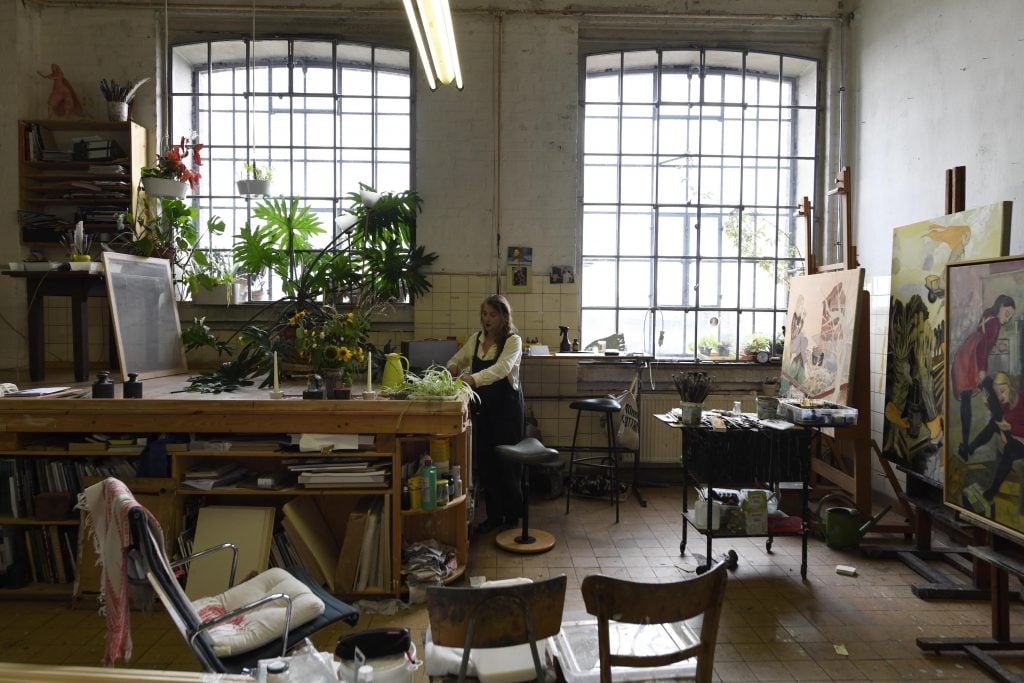 Rosa Loy , a middle aged white woman with long hair stands at an easel in an art studio.