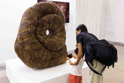 A child takes a photo of a large sculpture with a smartphone as his mother holds his arms. 