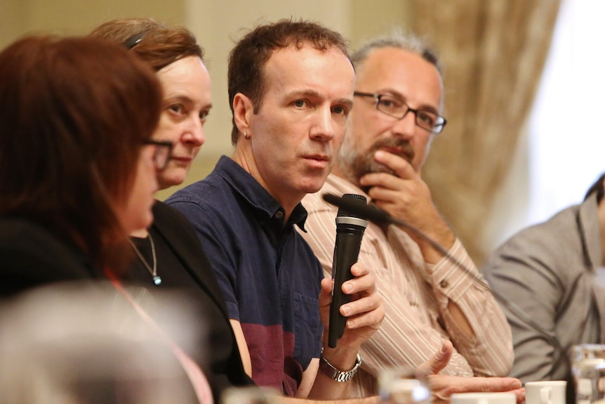 Man sitting at a table holds a microphone and speaks as other men and women watch on.