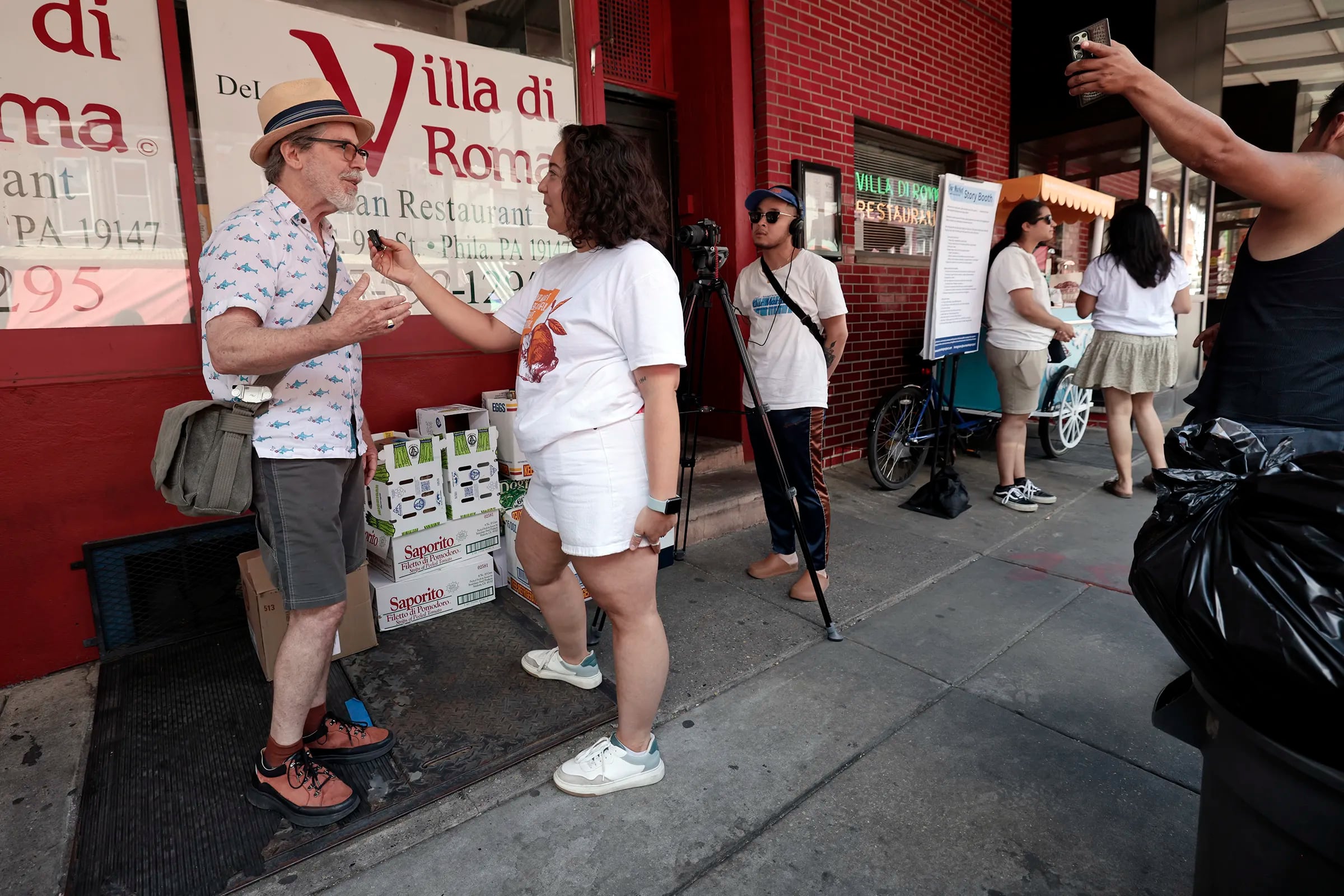 Stuart Rome of Philadelphia (left) is filmed while being interviewed by Our Market project manager Sophia Terry in the Italian Market in Philadelphia on Saturday, June 15, 2024. Local artist Michelle Angela Ortiz is collecting stories of the immigrant/migrant vendors, business owners, and neighbors that work and reside in the 9th Street Market for a community building public art project titled Our Market.