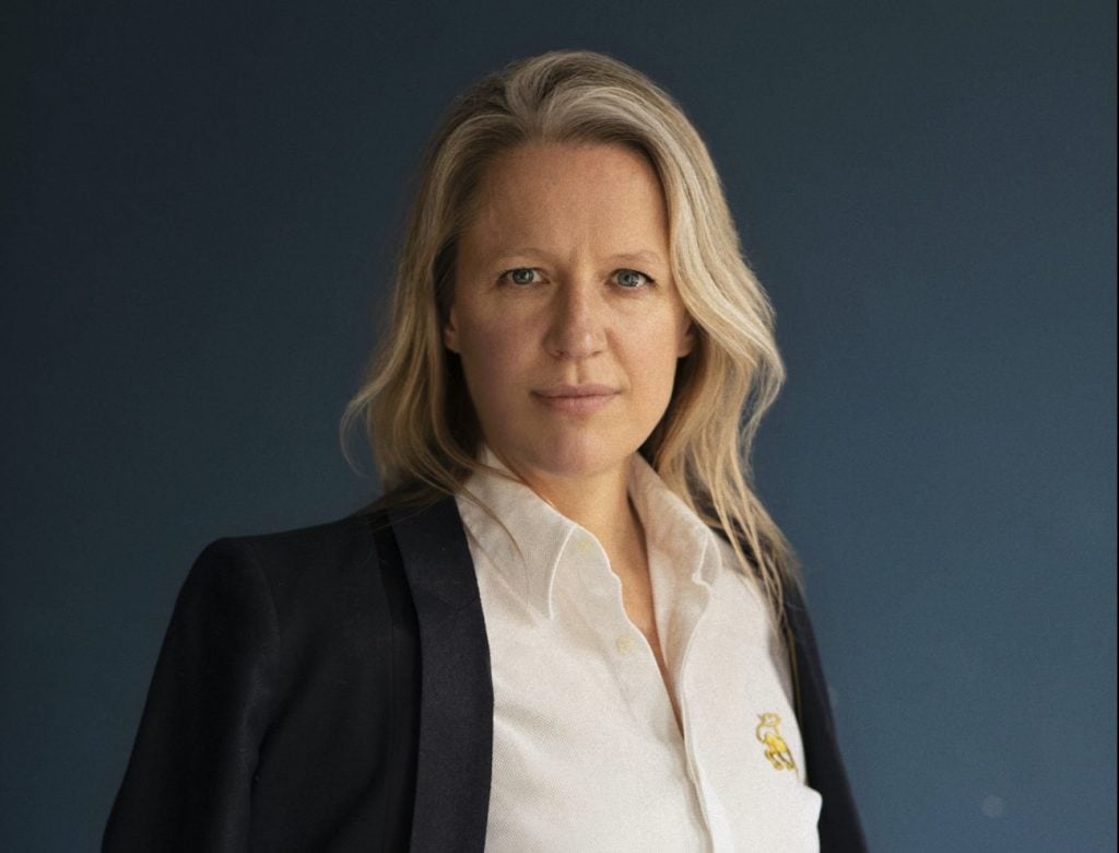 a woman in a white shirt and blazer stands against a blue wall