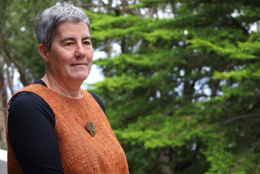 A woman stands slightly side on to the camera, with an out-of-focus pine tree in the background