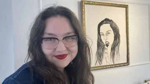 Lara King/BBC A woman with long dark hair, nose ring and rectangle glasses, wearing red lipstick. She is standing in front of a gold-framed sketch of a woman, which is in the background