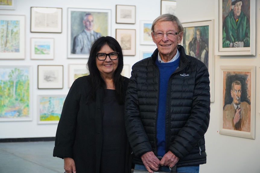 Two people, an older man and middle-aged woman, stand in an art gallery surrounded by portraits. 