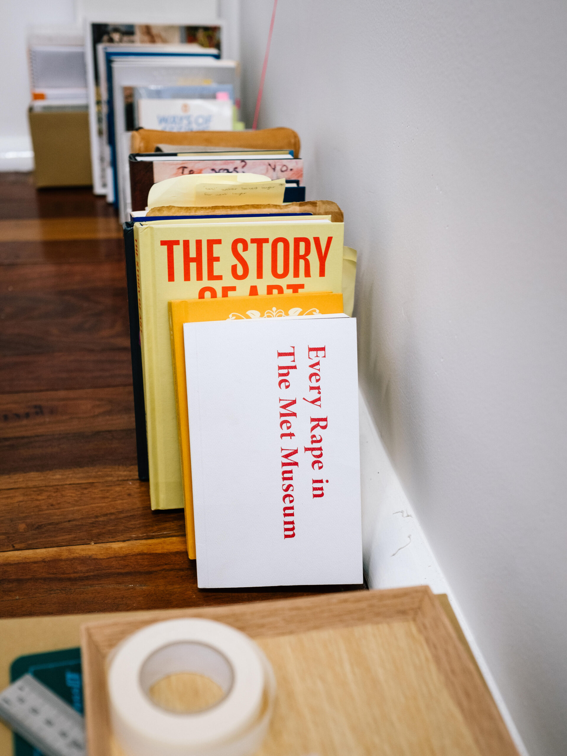 Books leaning against an art studio wall. The titled of the first is 'Every Rape in The Met Museum'