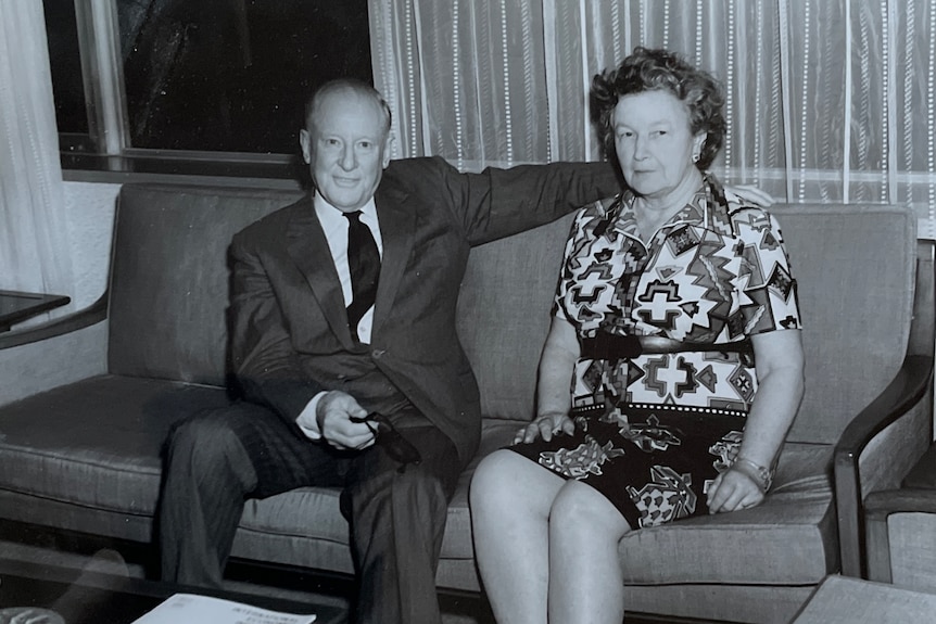 Bl;ack and white photo of a man and a woman sitting on a couch, the man's arm extended along the back of it.