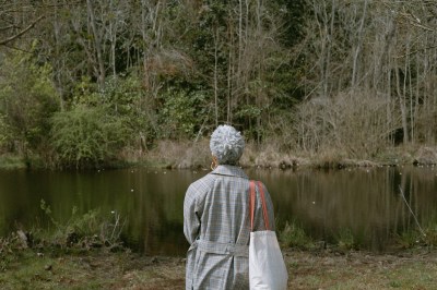 A woman with white-gray textured hair stares at a creek. She wears a patterned trench coat and carries a tote bag with orange straps. 