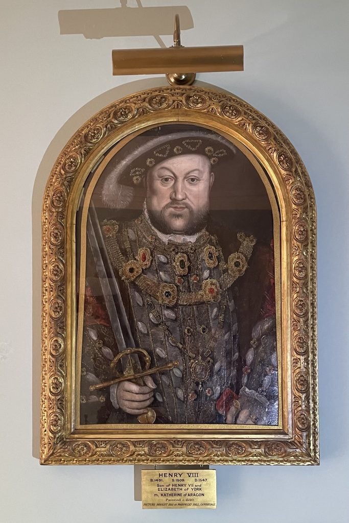 A framed portrait of Henry VIII displayed on a wall, featuring him in ornate royal attire with intricate jewelry and a feathered hat. He holds a sword, symbolizing his authority. The portrait is set within an elaborate gold frame with a plaque below that reads: 