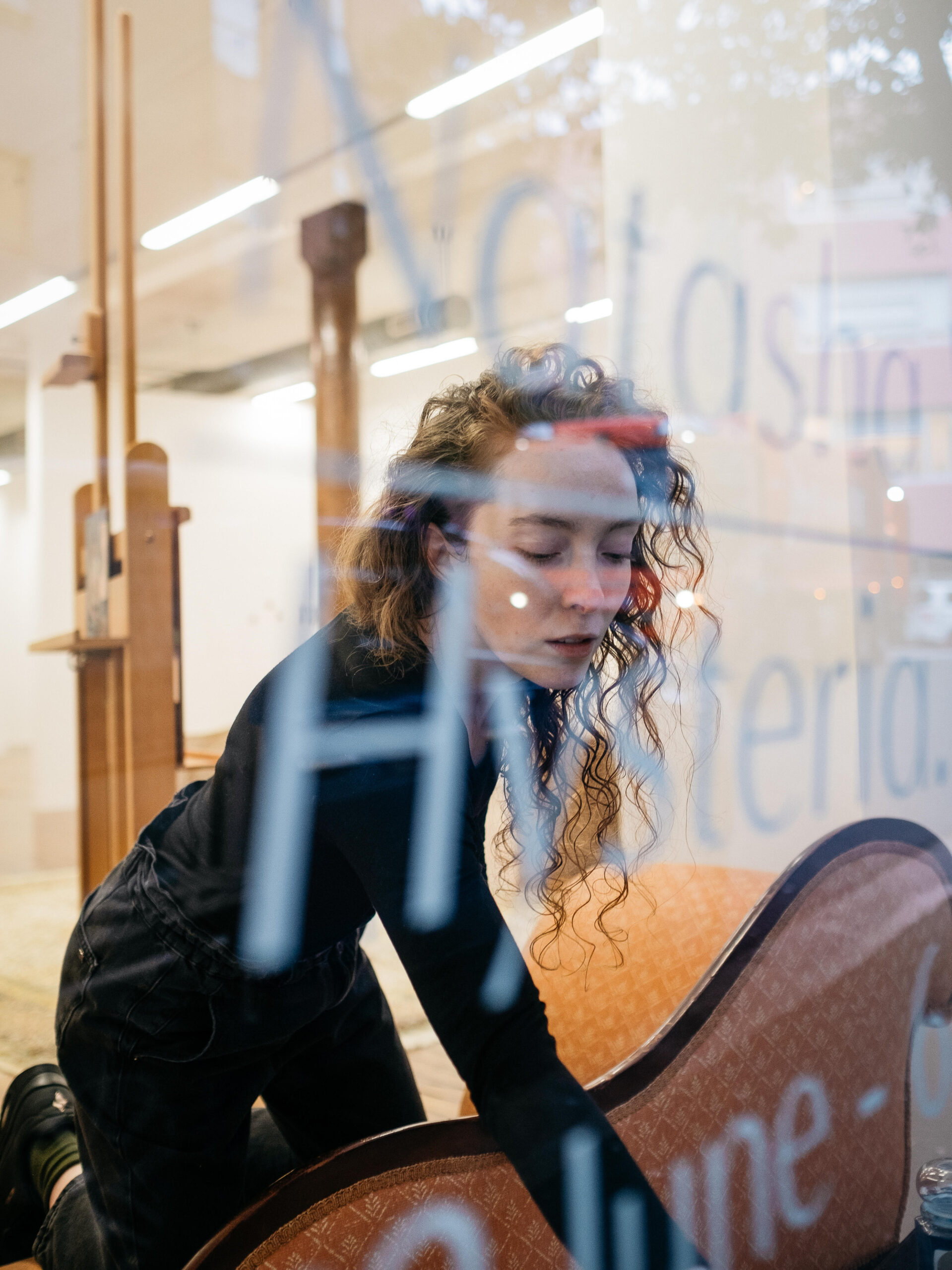 Photo portrait of the artist through the gallery window. She is reaching for something on the windowsill 