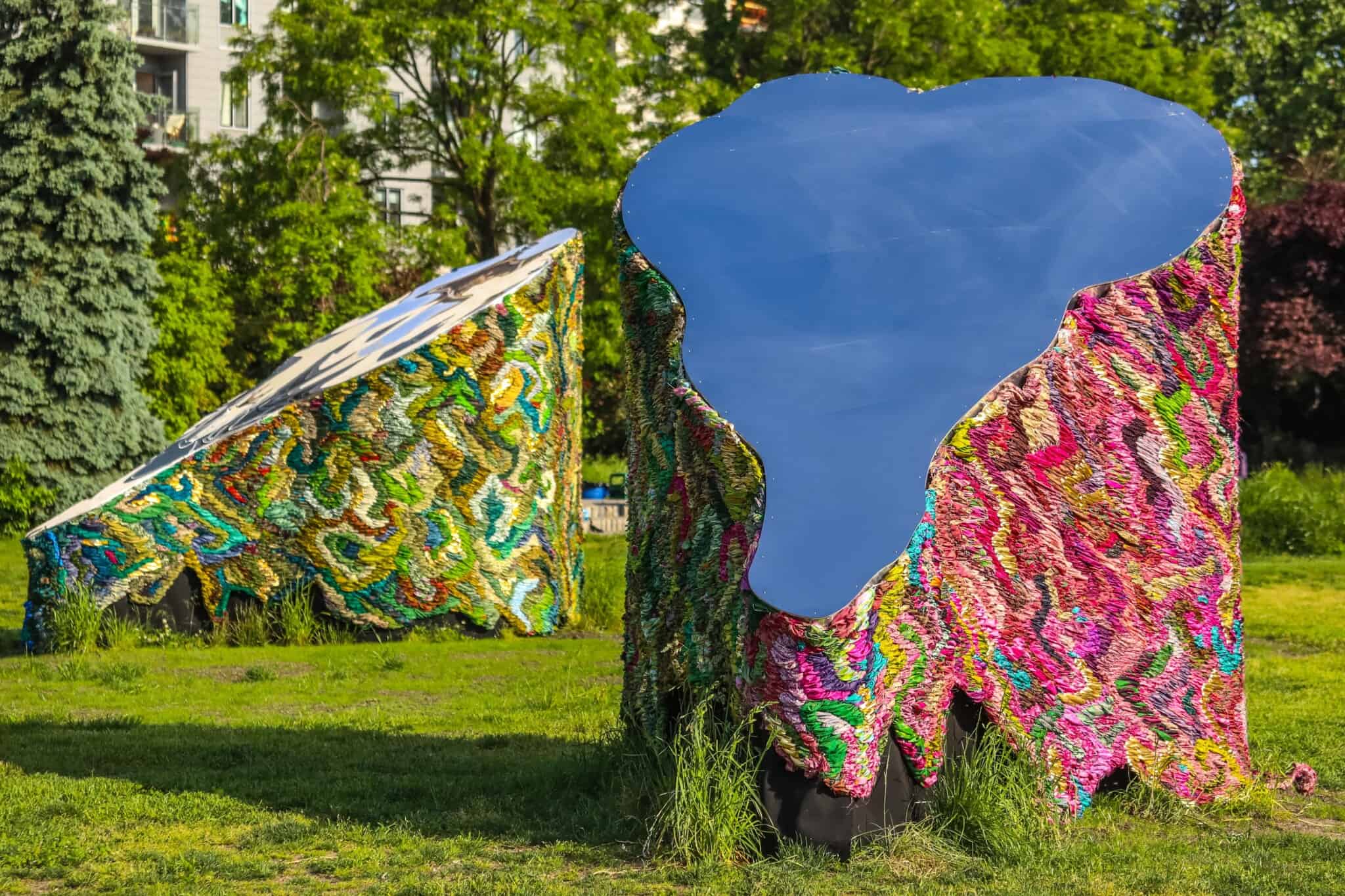 Two large pieces of art stand in a park, featuring textured fabric and reflective surfaces.