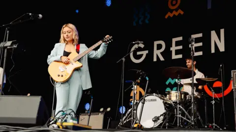 Nici Eberl Gillie Rowland, holding a guitar and wearing a blue suit, performs at Green Man