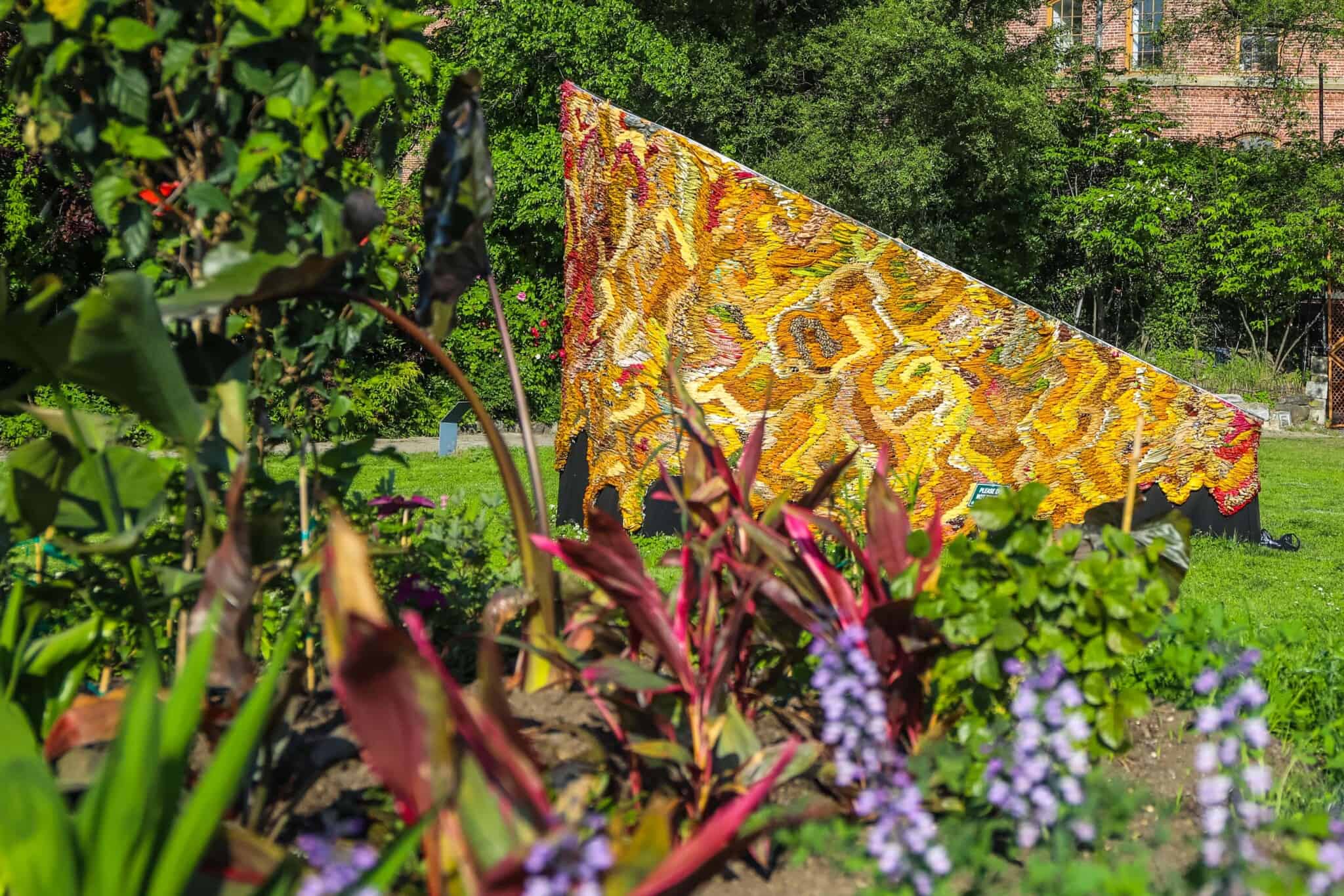 A piece of textured art, in yellow, golds, and browns, stands in a park, behind some flowers.