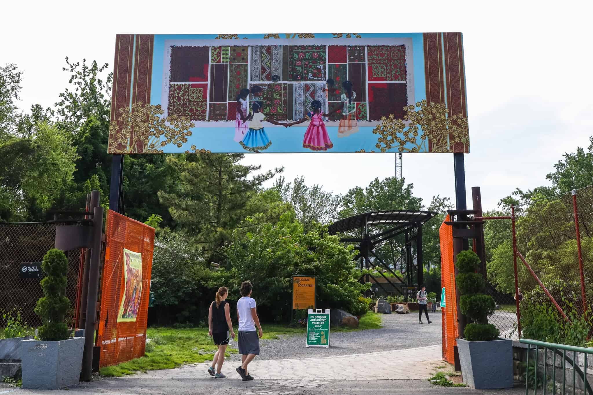 Two people walk through a gate into a park, with a mural featuring dark-skinned girls holding hands in a circle above them.