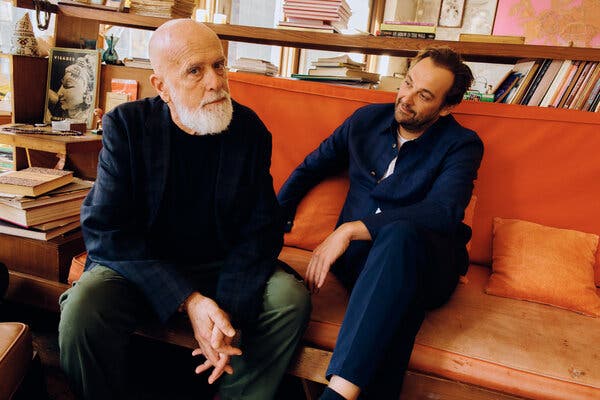 Two men, one wearing a blue buttoned shirt, the other with a bald head, white beard and a dark blazer on, sit on an orange couch in front of bookshelves.