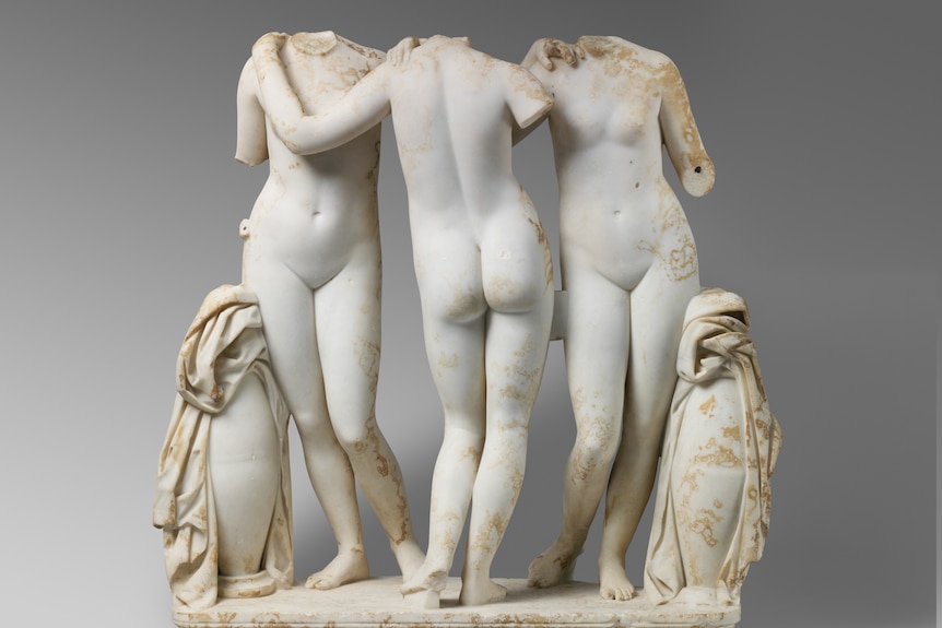 A marle statue of three headless female figures, standing nude, side by side, with arms on one another. 