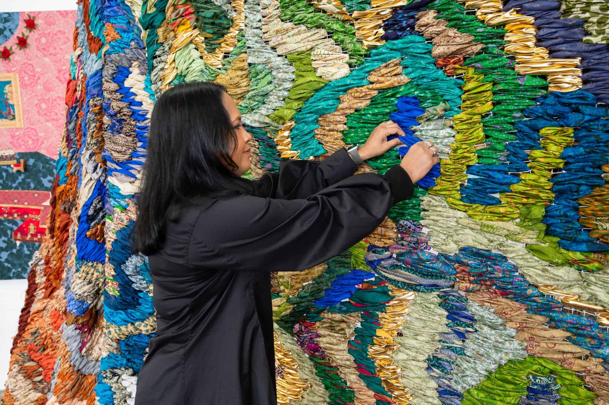 A south Asian woman, in black, inserts some fabric into a colorful wall of textured fabric.