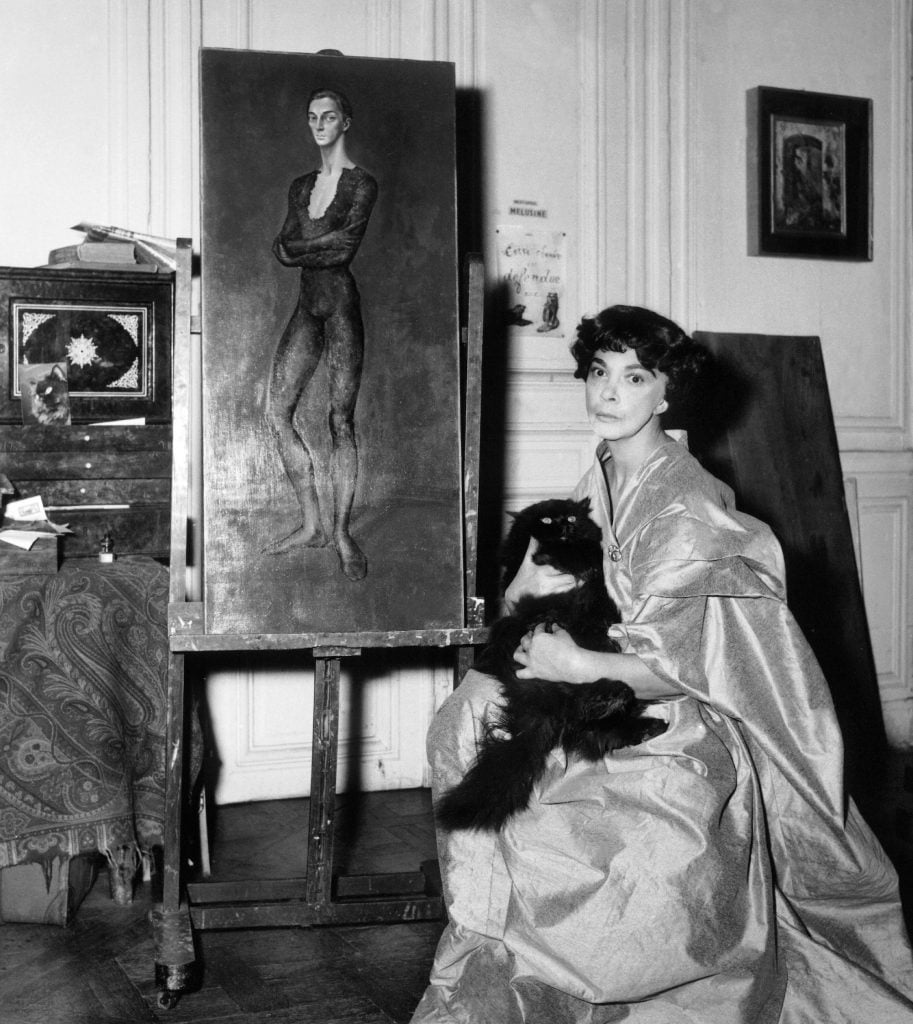 a black-and-white photograph of the artist leonor fini in a draping satiny gown and holding her fluffy black cat while she sits beside on of her paintings