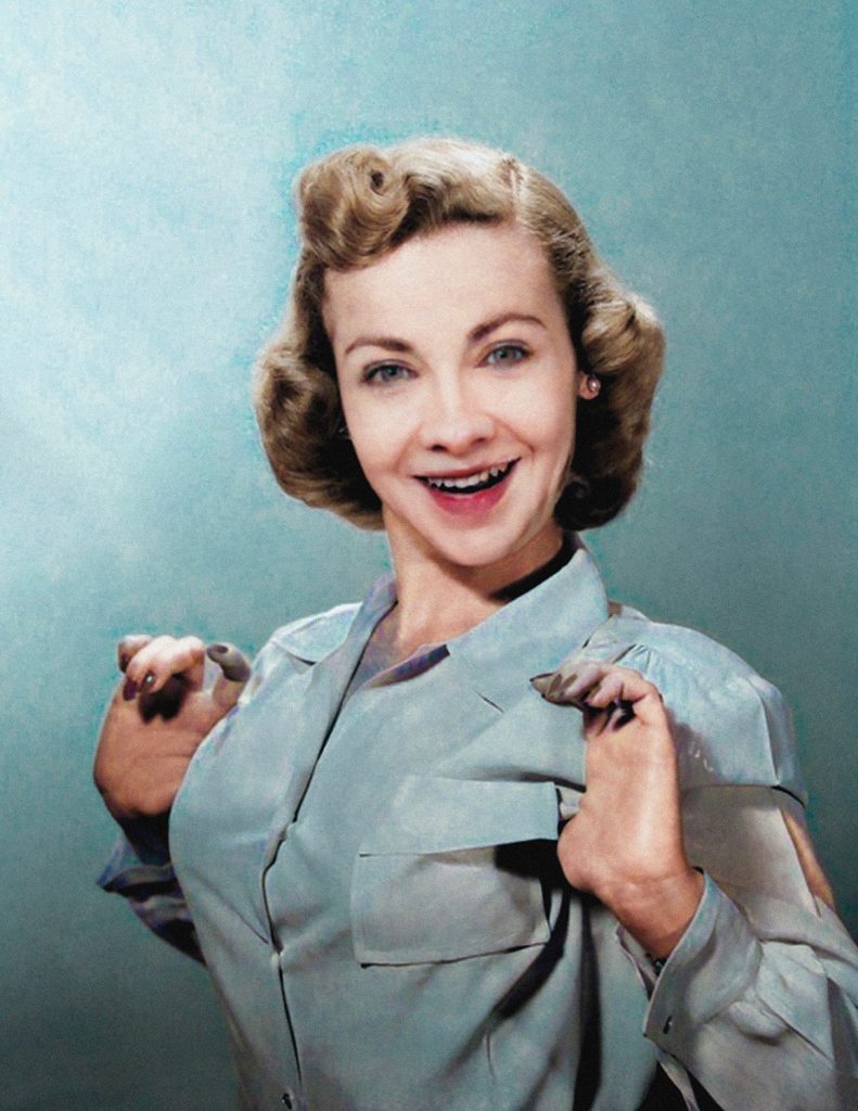 Portrait of a woman with a 1950s hairstyle and blue dress on a blue background smiling at the camera with her hands raised up to her chest with palms outward, found on the Artnet Gallery Network.