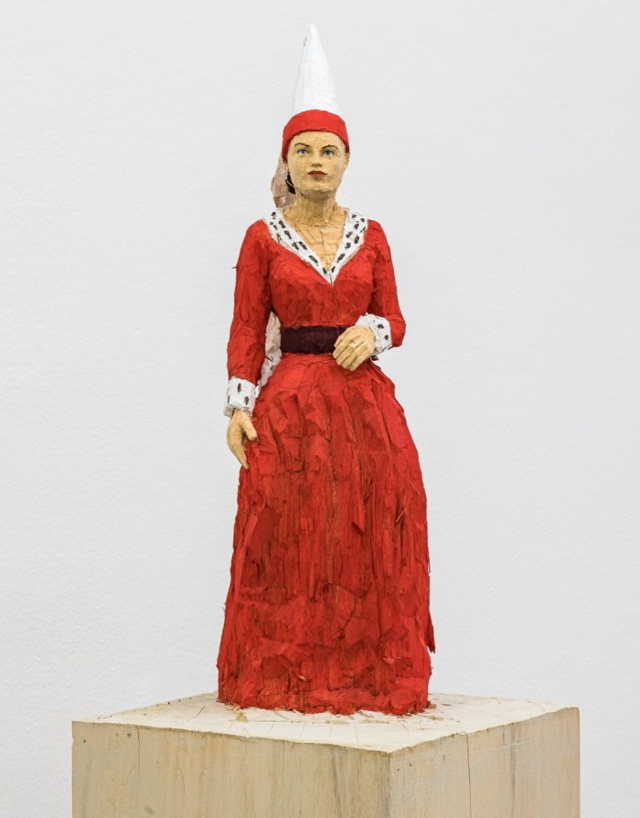 Roughly carved wooden sculpture of a woman in a medieval red dress with a white conical hat