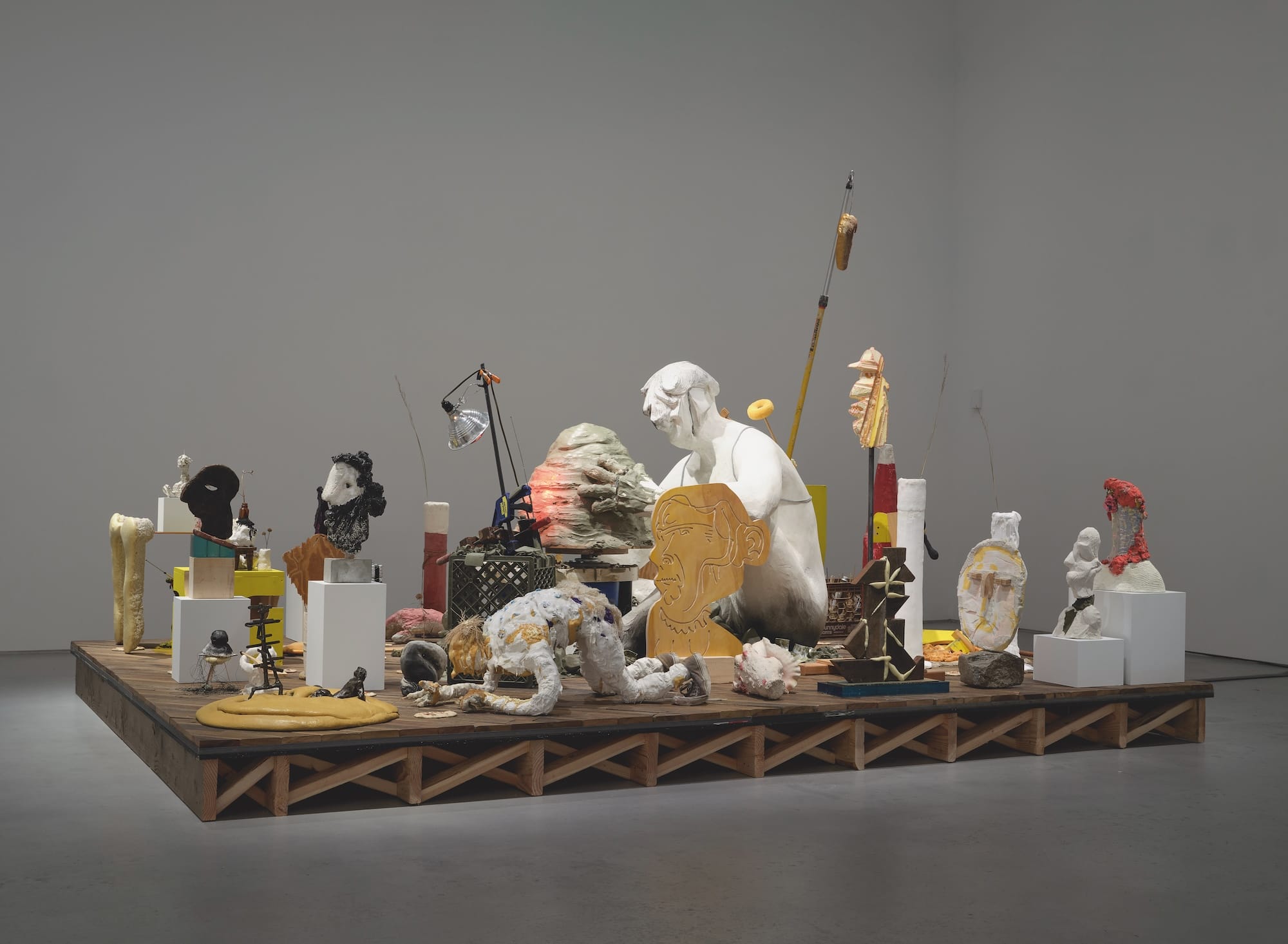 a mixed-media sculpture by Nicole Eisenman depicting abstract figures on a wooden platform in a muted gallery space