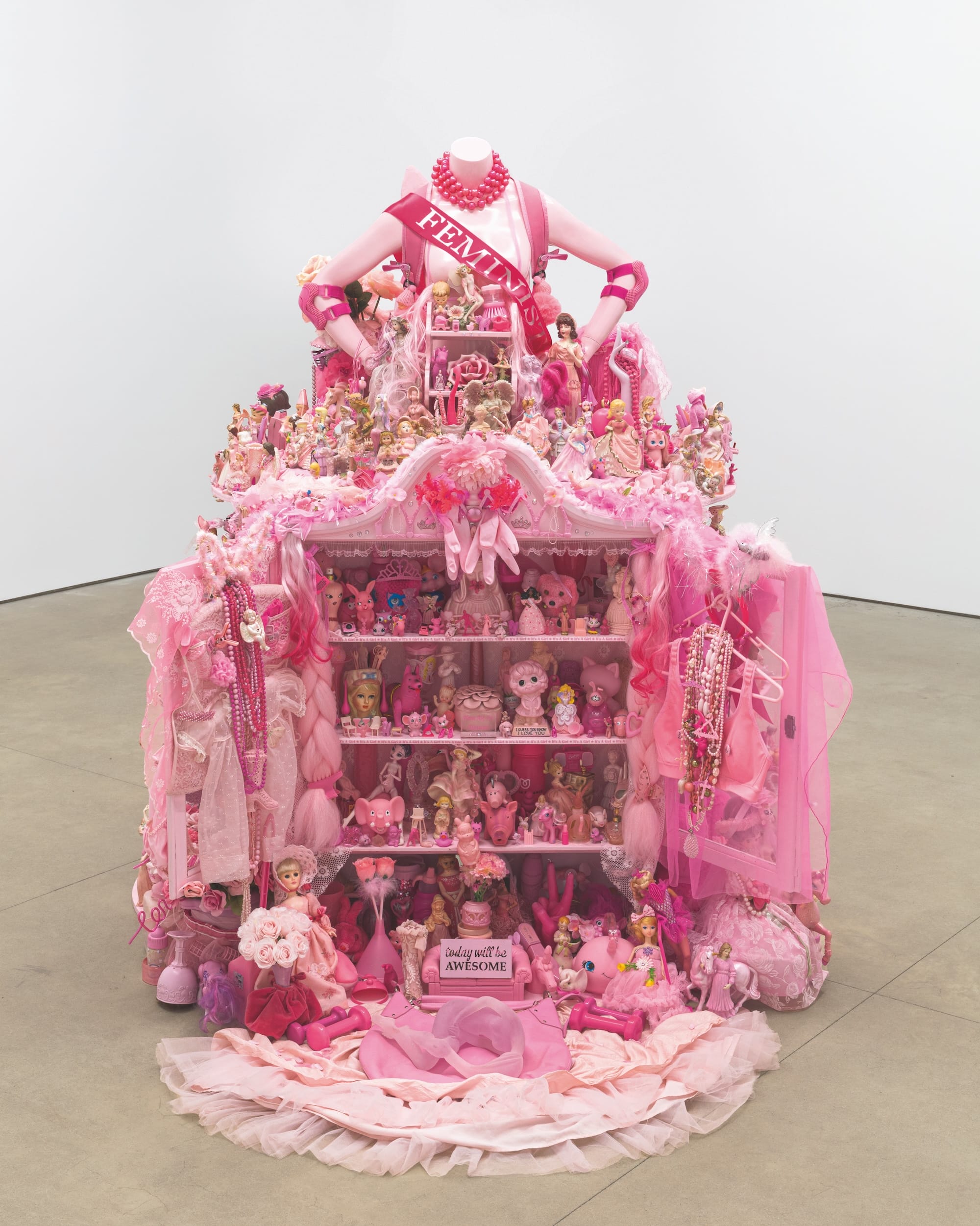 a sculpture made of all-pink found objects, topped with the torso of a female mannequin that appears to be opening a huge skirt to reveal shelves of objects and a sign that reads 