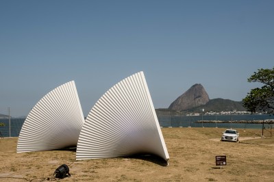 Two white sculptures that resemble fanned out paper are installed on sand in front of a harbor. 